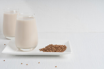 Gluten-free and lactose-free healthy buckwheat milk full of vitamins, minerals and antioxidants served in tall drinking glass with raw grains on plate on white wooden background. Image with copy space