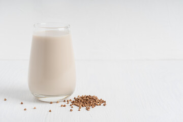 Gluten-free and lactose-free healthy buckwheat milk full of vitamins, minerals and antioxidants served in tall drinking glass with raw grains on white wooden background. Image with copy space