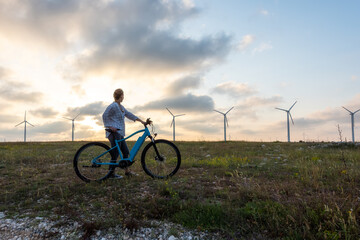 Woman with a bike in the nature / A woman with an electric bike enjoys the sunset against the backdrop of wind turbines. Green energy, sustainable alternative electricity, no pollution environment.