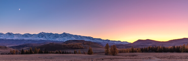 Beautiful panorama of a valley full of trees and white snowy mountains in the background. Sky is blue, orange, pink, and purple. Blue hour. Fall time. Sunset. Altai mountains, Russia