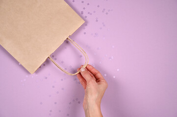Close up female hold in hand empty craft paper gift bag for purchases after shopping on pastel violet background. Packaging template mockup. Delivery concept advertising mock