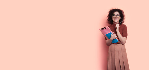 Charming curly haired student is smiling at camera through glasses wearing a dress on a studio wall with free space