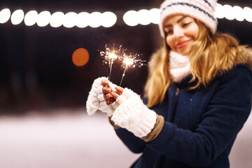 Female hand holding a burning sparkler firework bengal light.  Young woman celebrating holding sparkles in the winter forest. Festive garland lights. Christmas, New year party.