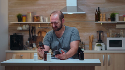 Man with headache holding pills bottle sitting in the kitchen. Stressed tired unhappy worried person suffering of migraine, depression, disease and anxiety feeling exhausted with dizziness symptoms.