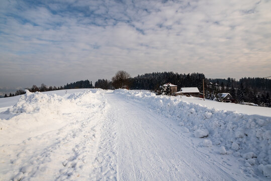 winter scenery with snow covered road, few houses, smaller hills and blue sky with clouda above Bukovec village in Czech republic