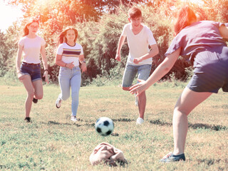 Happy teenage girls and guy playing football on green lawn in summer city park
