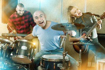 Expressive smiling glad friendly drummer with his bandmates practicing in rehearsal room