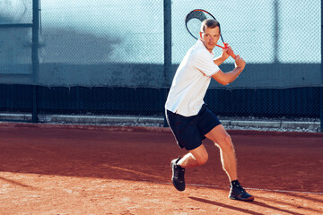 Young man plays tennis outdoors on tennis court in the morning