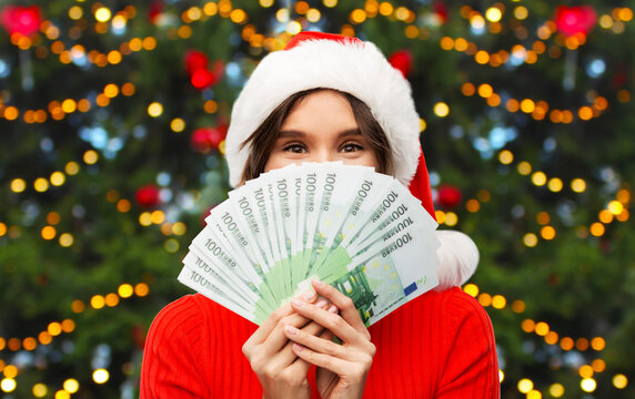 winter holidays and finance concept - happy smiling young woman in santa helper hat holding euro money banknotes over christmas tree lights on background