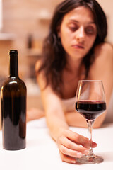 Glass of wine in focus while woman is being lonely, hangover and depressed. Unhappy person disease and anxiety feeling exhausted with having alcoholism problems.