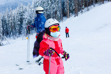 Fototapeta na wymiar Portrait of a little girl skier in medical mask during COVID-19 coronavirus on a snowy mountain at a ski resort