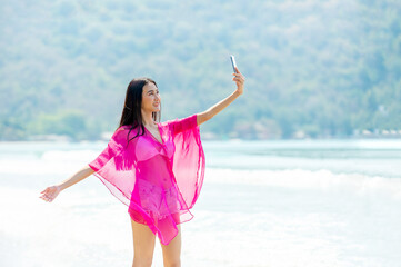 Happy young beautiful Asian woman walking on the beach and using smartphone taking selfie in summer sunny day. Pretty sexy girl enjoy and having fun with outdoor lifestyle and summer holiday vacation.