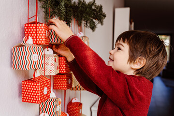 Little happy curious child opening first gift of handmade advent calendar hanging on wall....