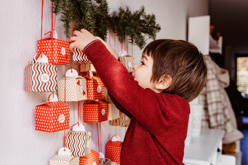 Little happy curious child opening first gift of handmade advent calendar hanging on wall. Sustainable Christmas. Celebrating at home, childhood.