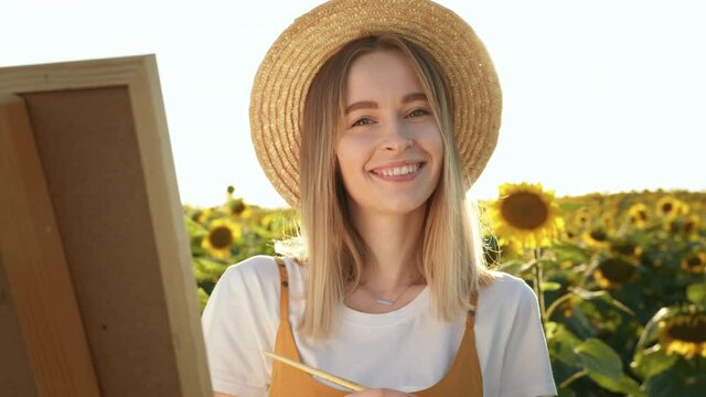 A woman is standing in a field of sunflowers and drawing a picture. She is looking at the camera and smiling. Close-up shooting. 4K
