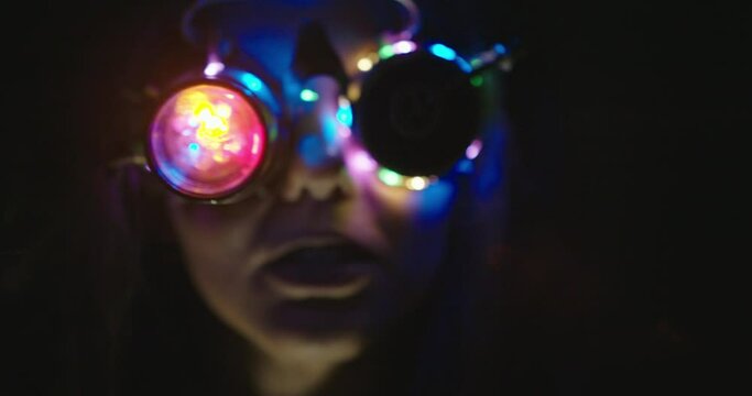 Face of a modern hipster young woman wearing funky goggles with flashing colorful lights on the rims in a dark shadowy atmospheric environment moving seductively in front of the camera