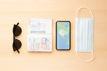 Flat lay, travel concept during the Covid-19 outbreak, a face mask, a passport, sunglasses and a smartphone with a World Map are arranged on a wooden table. 