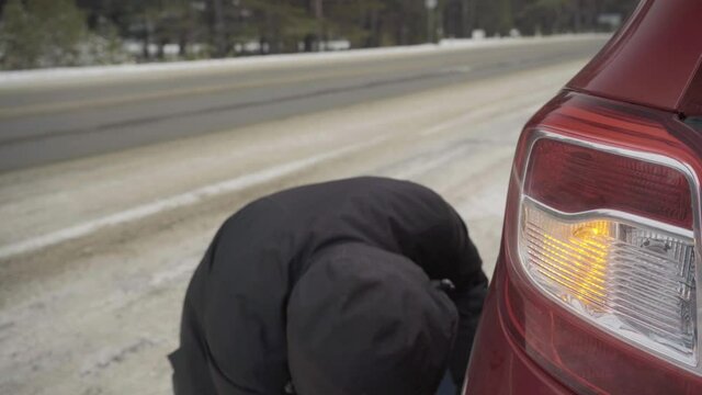 changing a wheel on the road in winter. car breakdown on the road. a man in a black jacket unscrews or spins a wheel. the headlight of the red car flashes. do it yourself car repair.