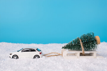 Christmas celebration concept. Close up photo of toy mini car carrying white wooden sledge with little christmas tree on isolated on blue background