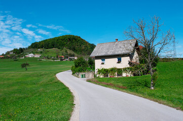 Fototapeta na wymiar House near road going down the hill through field. Blue sky and green grass. Slovenia valley.
