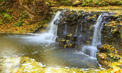 
A small waterfall in the forest flows over the black stones in autumn. Long exposure.