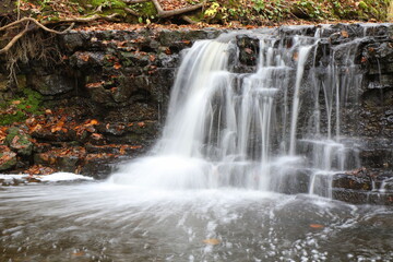 
A small waterfall in the forest flows over the black stones in autumn. Long exposure.