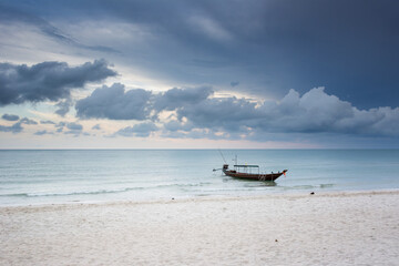 One long tail boat moored near the shore in Thailand. A dark sky like a storm is coming, low clouds on the horizon. Calm before the storm. White sand, nobody, just a boat. Bottle Beach, Koh Phangan