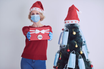 Woman with gift box present, decorating christmas tree with medical masks during virus pandemic 2020 / 2021.