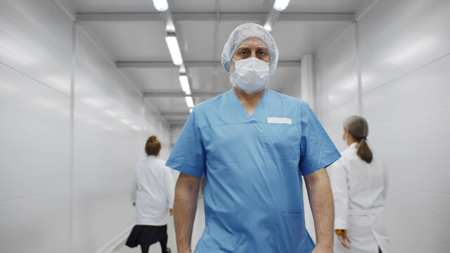 Senior Male Surgeon In Uniform, Cap And Facial Mask Walking Along Corridor In Clinic