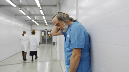Stressed tired aged surgeon leaning on wall in hospital corridor after difficult surgery