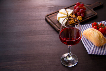 A glass of red wine, cheese, grapes, walnuts on a wooden cutting Board. Bread roll and cherry tomatoes on a napkin. Dark background. The concept of a romantic dinner. Top view. Copy space.