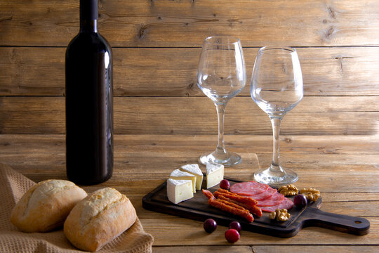 Two Wine Glasses And A Bottle Of Red Wine, Cheese, Meat Delicacies, Walnuts On A Wooden Cutting Board, Two Bread Rolls On A Napkin. The Concept Of A Romantic Dinner In A Restaurant Or At Home.