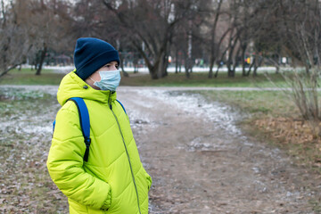 A boy in a bright yellow jacket walks in a city Park in autumn in a medical mask.