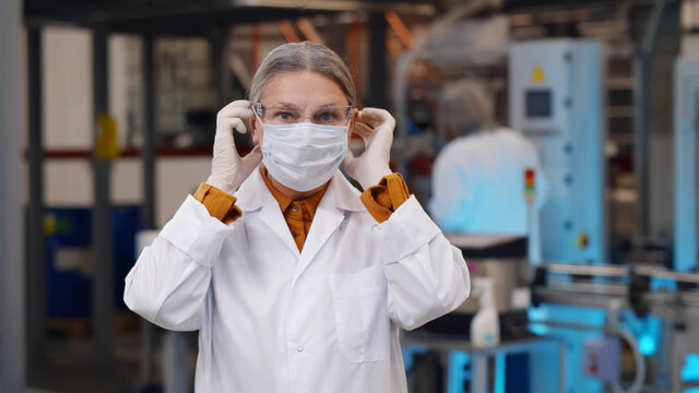 Portrait Of Mature Woman Scientist In Robe And Protective Glasses Putting On Safety Mask Standing At Plant