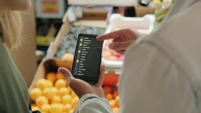 Tracking Over The Shoulder Shot Of Couple Looking At Shopping List On Mobile Phone And Chatting While Standing At Fruit Stand At Market