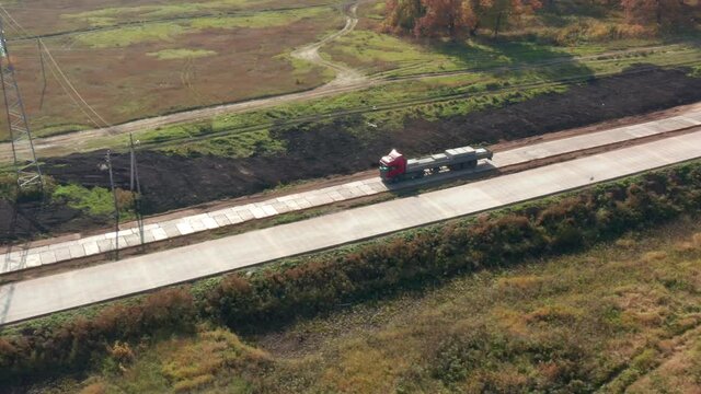 Aerial Follow Shot of RED Truck Moving Through Rural Area. Sun Shines. In the Background forest and other road are Seen. Red flatbed truck.