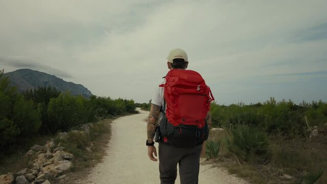 Camera follows young adult man with red backpack. Male traveller or hiker walk on empty gravel path or trail on cloudy summer day. Wanderlust travel inspiration for staycation ideas
