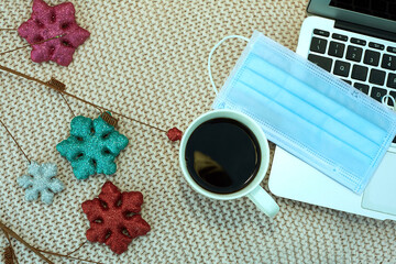 Mug with coffee, medical mask, laptop, New Year's symbols are located on a knitted blanket. Remote work concept during coronavirus infection.