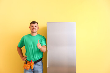 Worker of repair service showing thumb-up near fridge on color background