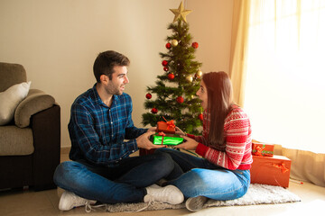 young couple smiling and surprised giving each other a gift with a christmas tree in the background