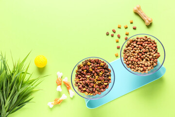 Bowls with dry pet food and grass on color background