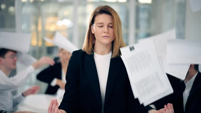 Calm blonde business woman dressed in classic black suit meditate in lotus position while office workers arguing and throwing papers to each other faces on business meeting at the background