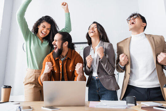 Excited hispanic office workers with yeah gesture standing near laptop at workplace