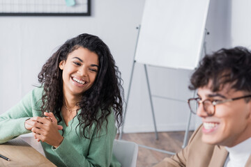 Fototapeta premium Curly hispanic woman laughing, while looking at co-worker sitting at desk in office on blurred foreground