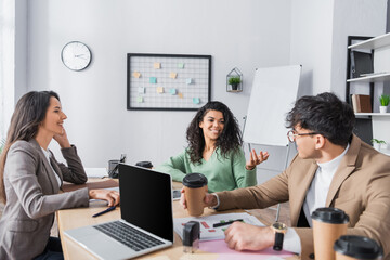 Smiling hispanic office workers looking at each other, while talking at workplace with device in...