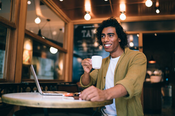 Happy prosperous young businessman in casual wear holding coffee cup enjoying break in cafe while blogging, smiling hipster guy freelancer satisfied with remote job and communication on laptop