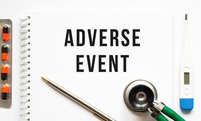 ADVERSE EVENT is written in a notebook on a white table next to pills and a stethoscope.