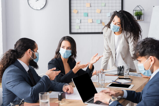 Hispanic businesswoman in medical mask looking at co-workers talking during business meeting in office