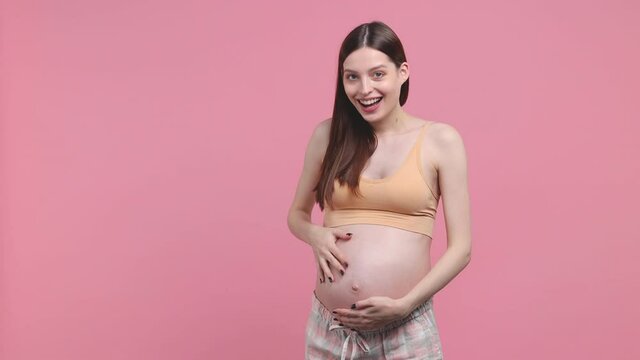 Surprised young pregnant woman future mom in basic top stroking keeping hands on belly stomach tummy with baby isolated on pastel pink background studio. Maternity family pregnancy gynecology concept