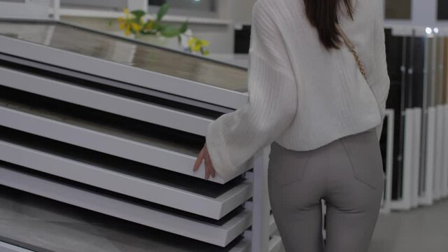 Young woman in white pullover walks along hardware store choosing countertop for living room table under electric light. Concept design work